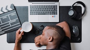 Man sleeping on desk with work items - Philanthropeak Coaching a man sitting at a desk with a laptop and headphones. Is Procrastination a Sign of ADHD? Understanding the Connection for Entrepreneurs