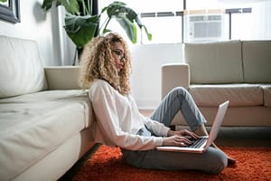 Woman working on laptop in comfortable living room - Philanthropeak Coaching woman sitting on floor and leaning on couch using laptop ADHD Coach
